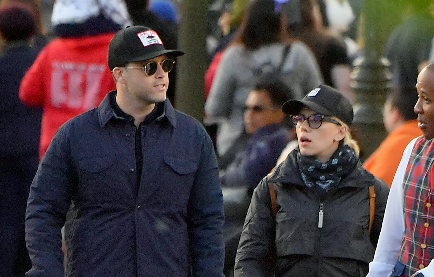 Colin Jost, in a baseball cap and sunglasses, walks alongside Scarlett Johannson who wears a baseball cap and coat.