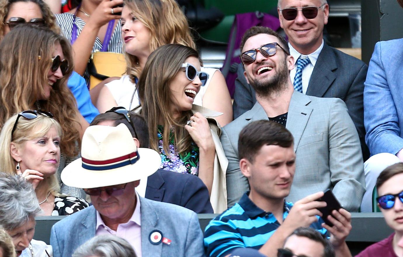 Jessica Biel, in a white jacket and patterned dress and sunglasses shares a laugh with Justin Timberlake who wears a grey jacket and black top. They’re in the stands at Wimbledon.