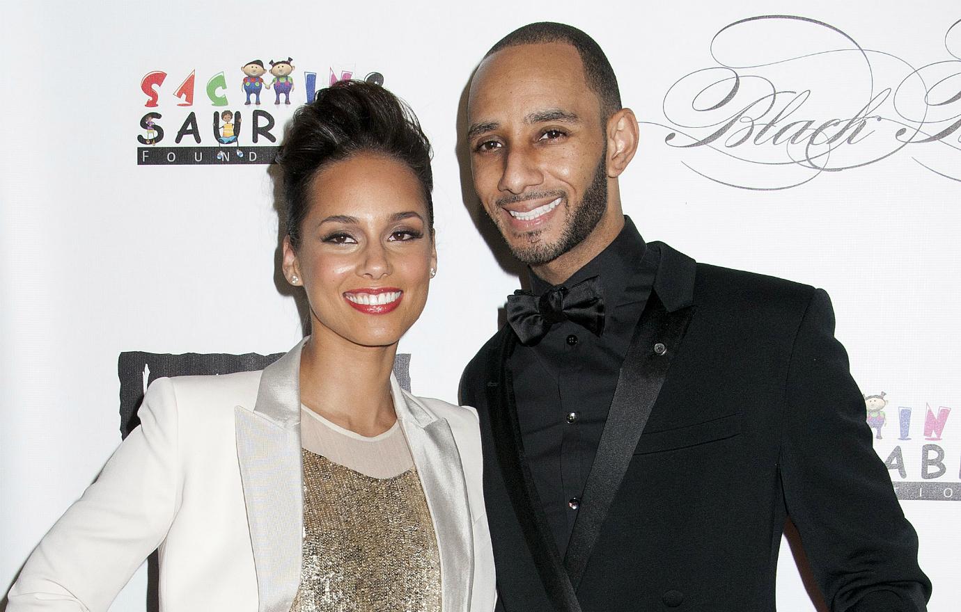 Alicia Keys wears a white jacket and glittery top and poses alongside Swizz Beatz, who wears a black suit, on the red carpet.