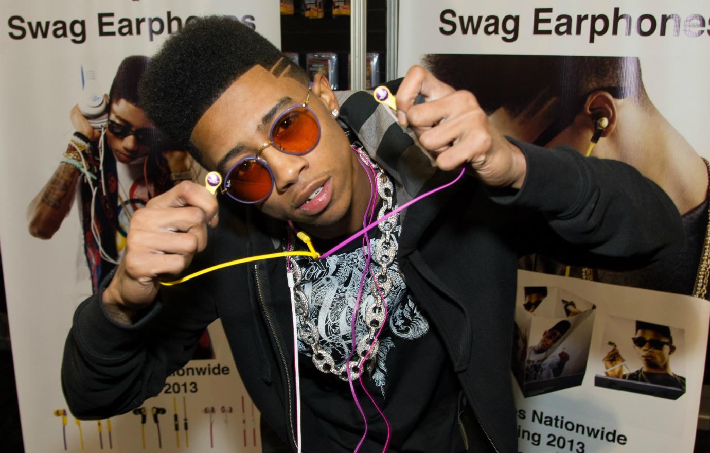 Lil Twist, in a black patterned shirt and sunglasses, holds earphones up for the camera.