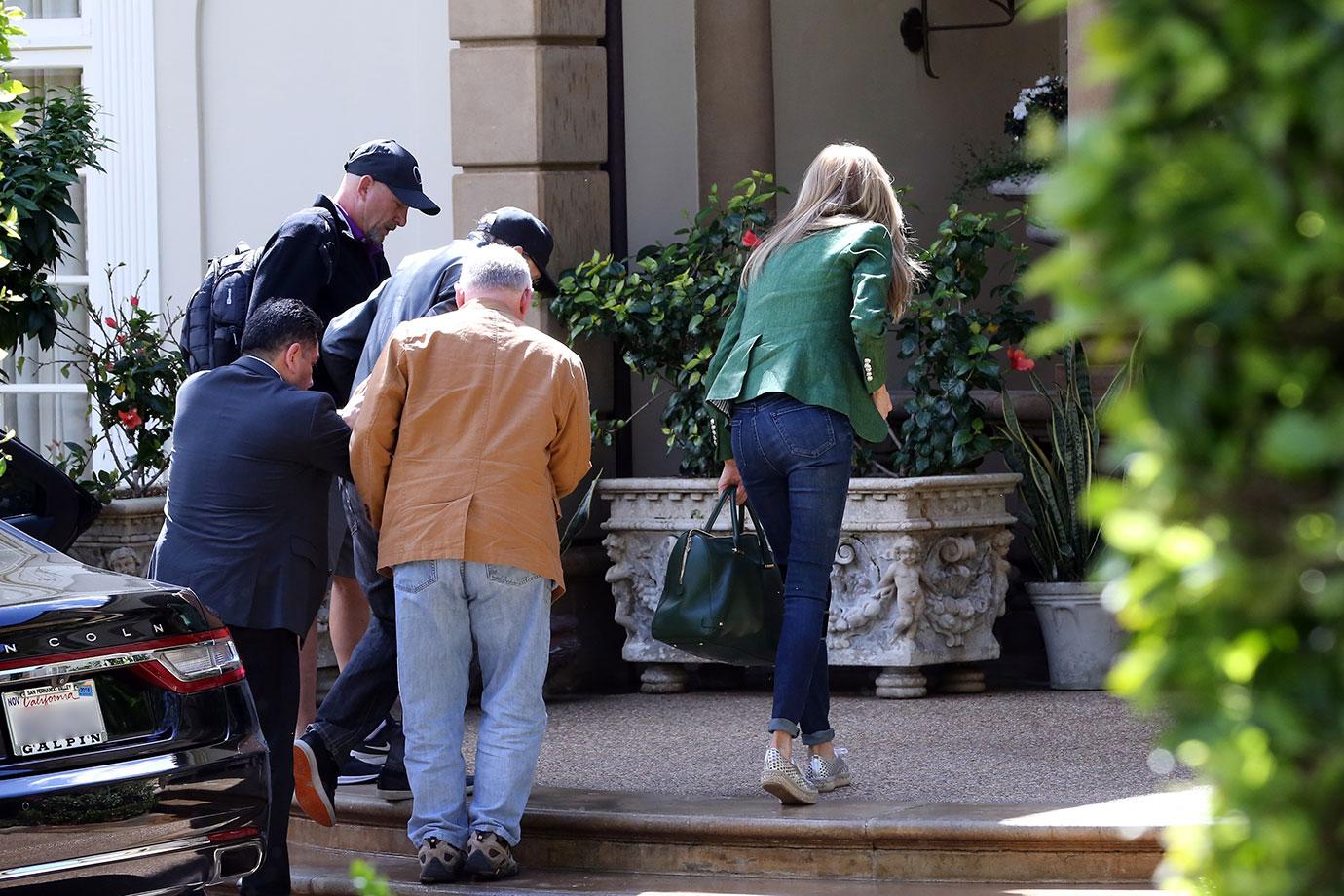 Larry King Wearing Baseball Cap Being Helped Out Of Car While Shawn Wearing a Green Jacket And Jeans Picks Up Purse and Goes Inside House