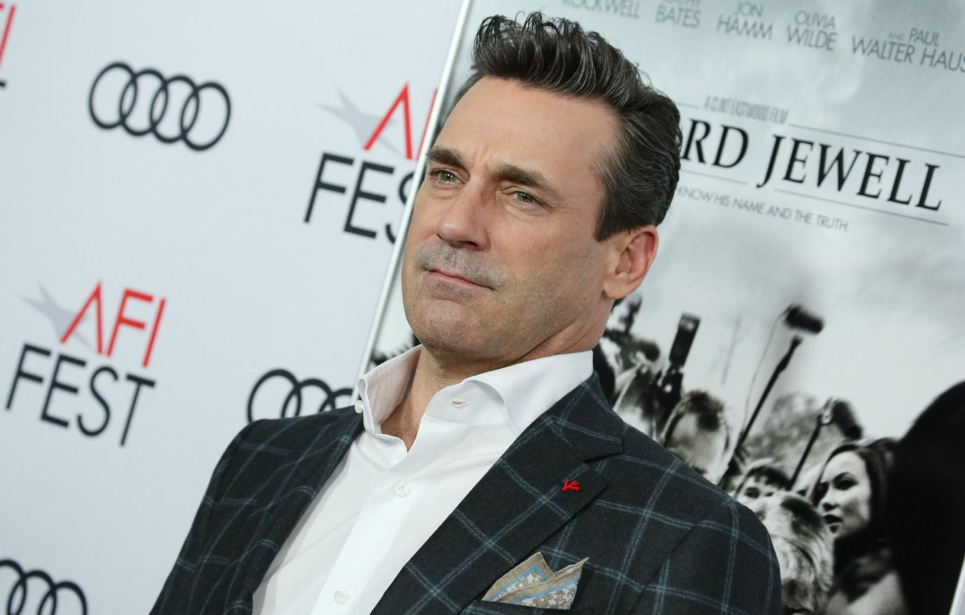 Jon Hamm, in a white shirt and check jacket, stands on the red carpet.