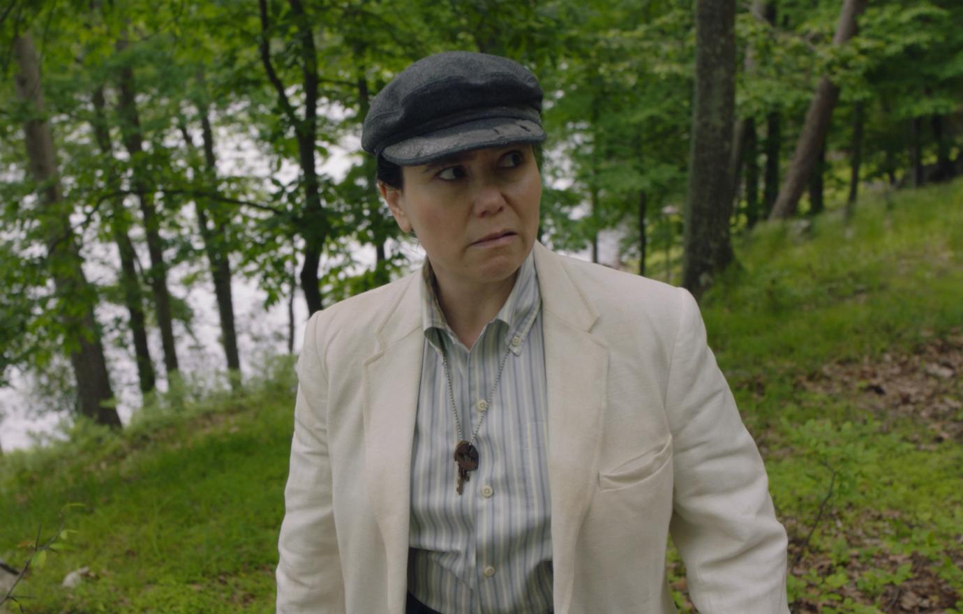 Alex Borstein, in character as Susie Myerson, wears a blue striped shirt, cream jacket, and dark hat.