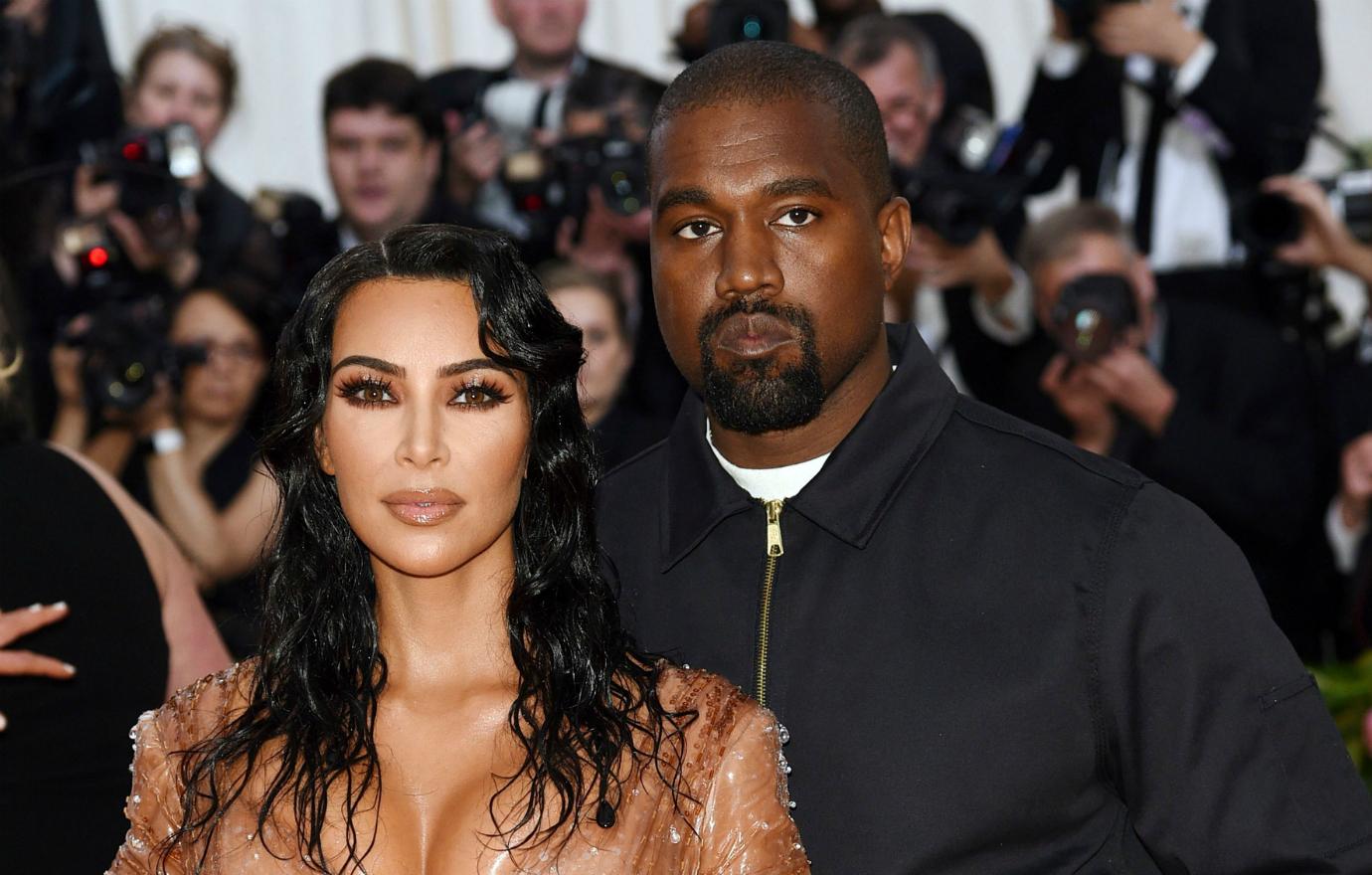 Kanye West, in a zip up top, poses next to Kim Kardashian who wears an orange dress.