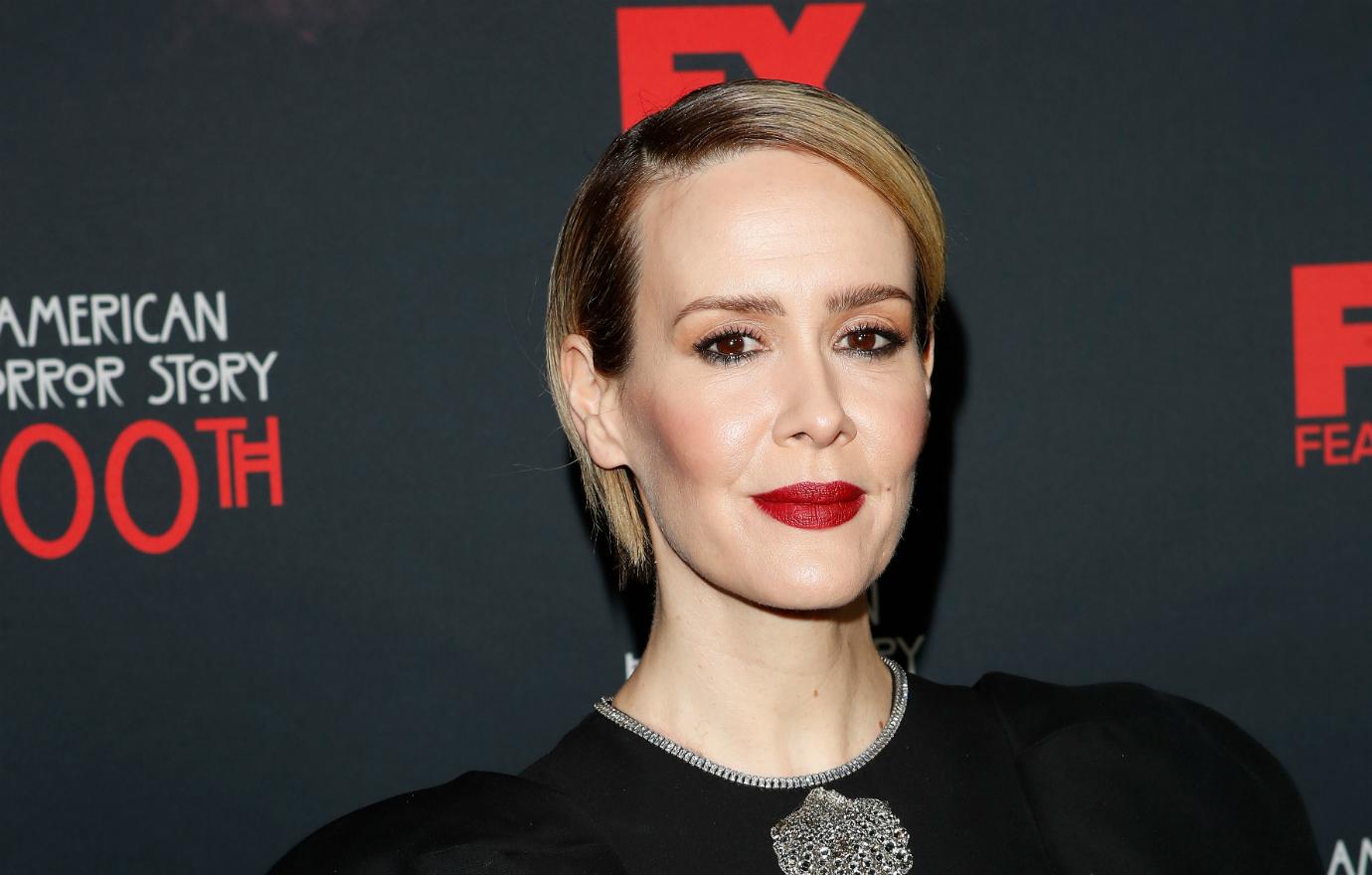 Sarah Paulson smiles at the camera on the red carpet. She wears a black dress.