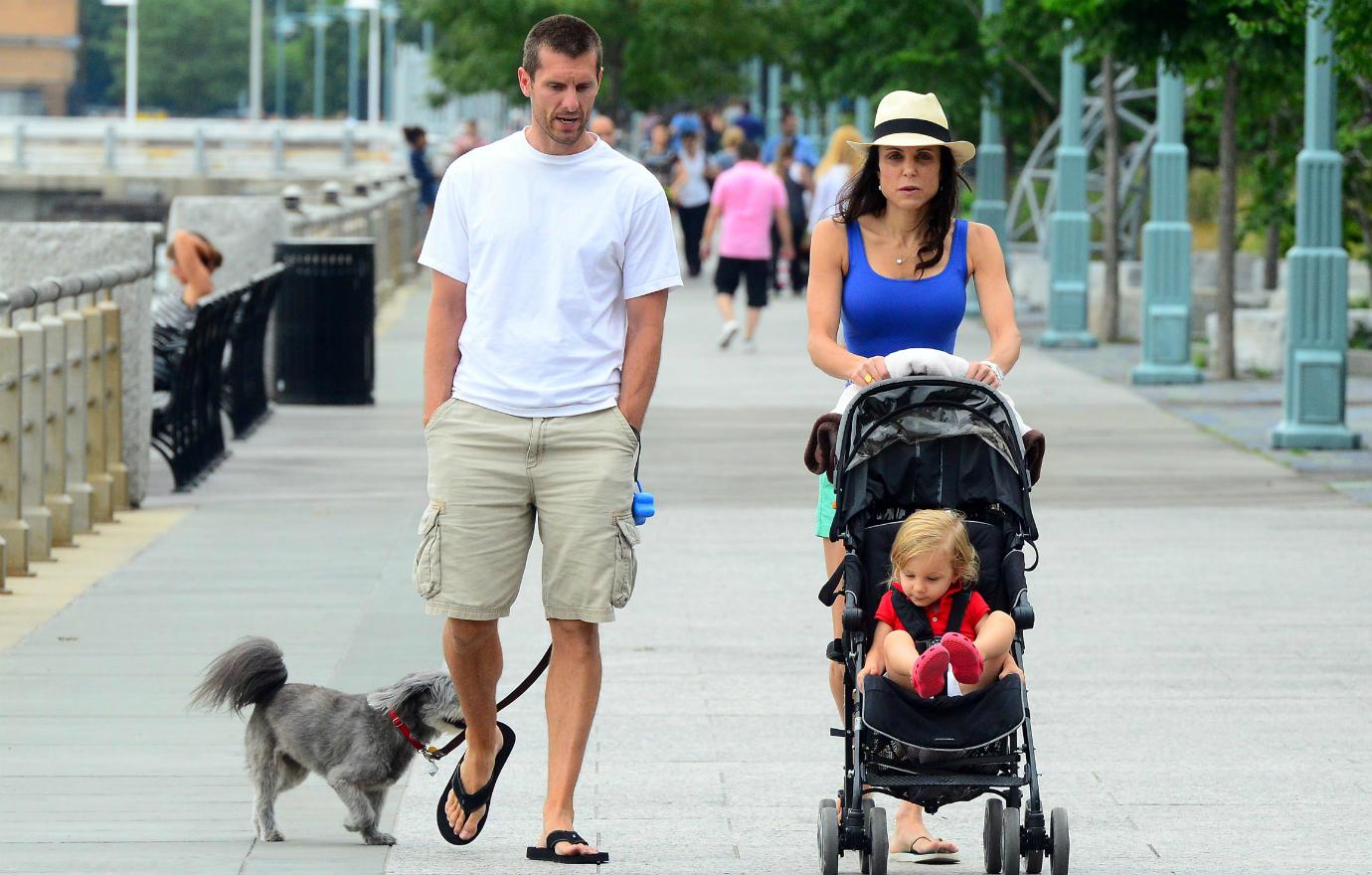 Jason Hoppy, in white top and shorts, walks alongside Bethenny Frankel, in blue, who pushes their child in their stroller.