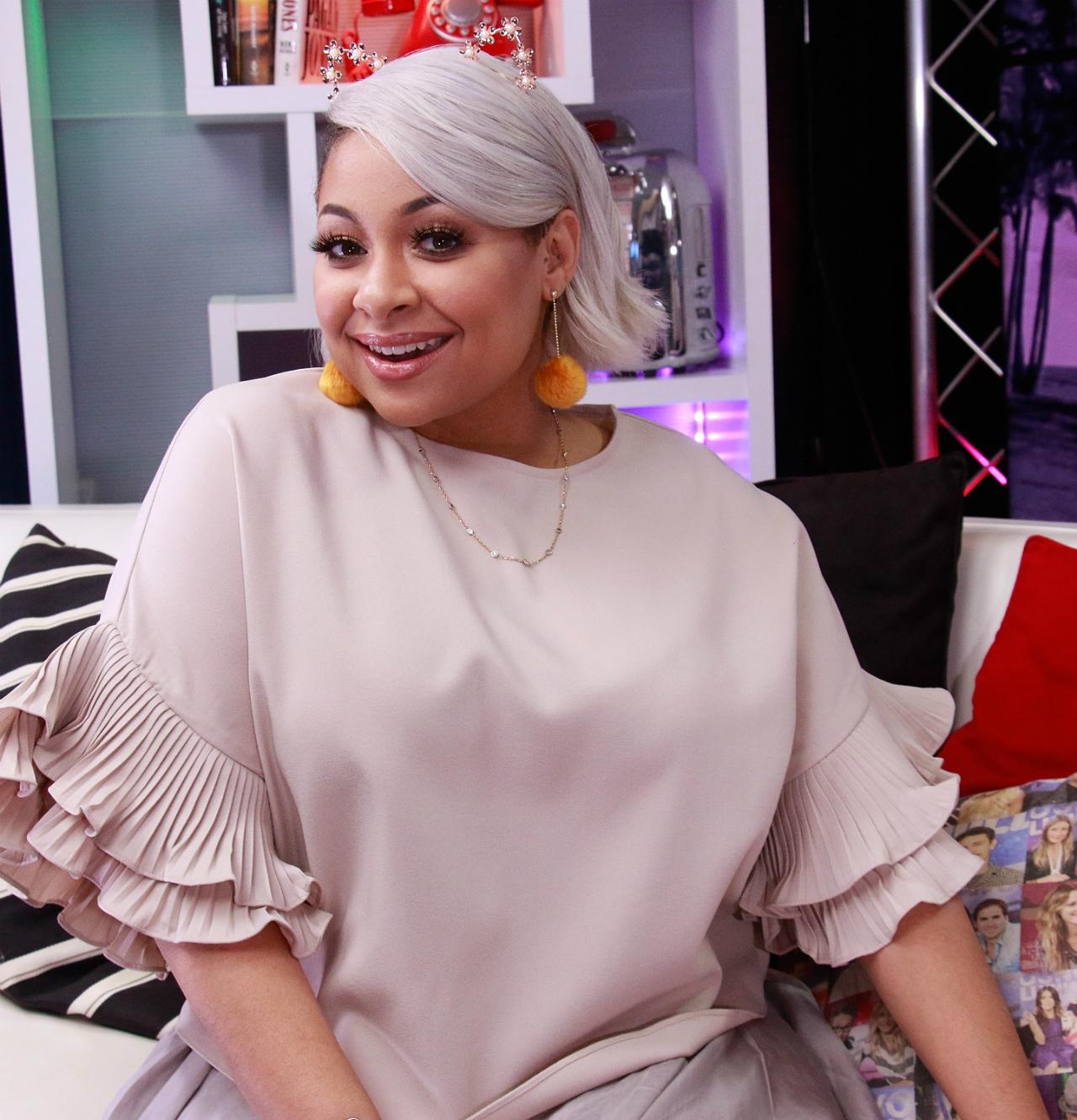 Raven Symone wears a blush pink shirt with short ruffled sleeves, brightly colored pom pom dangling earrings, and lavender hair.