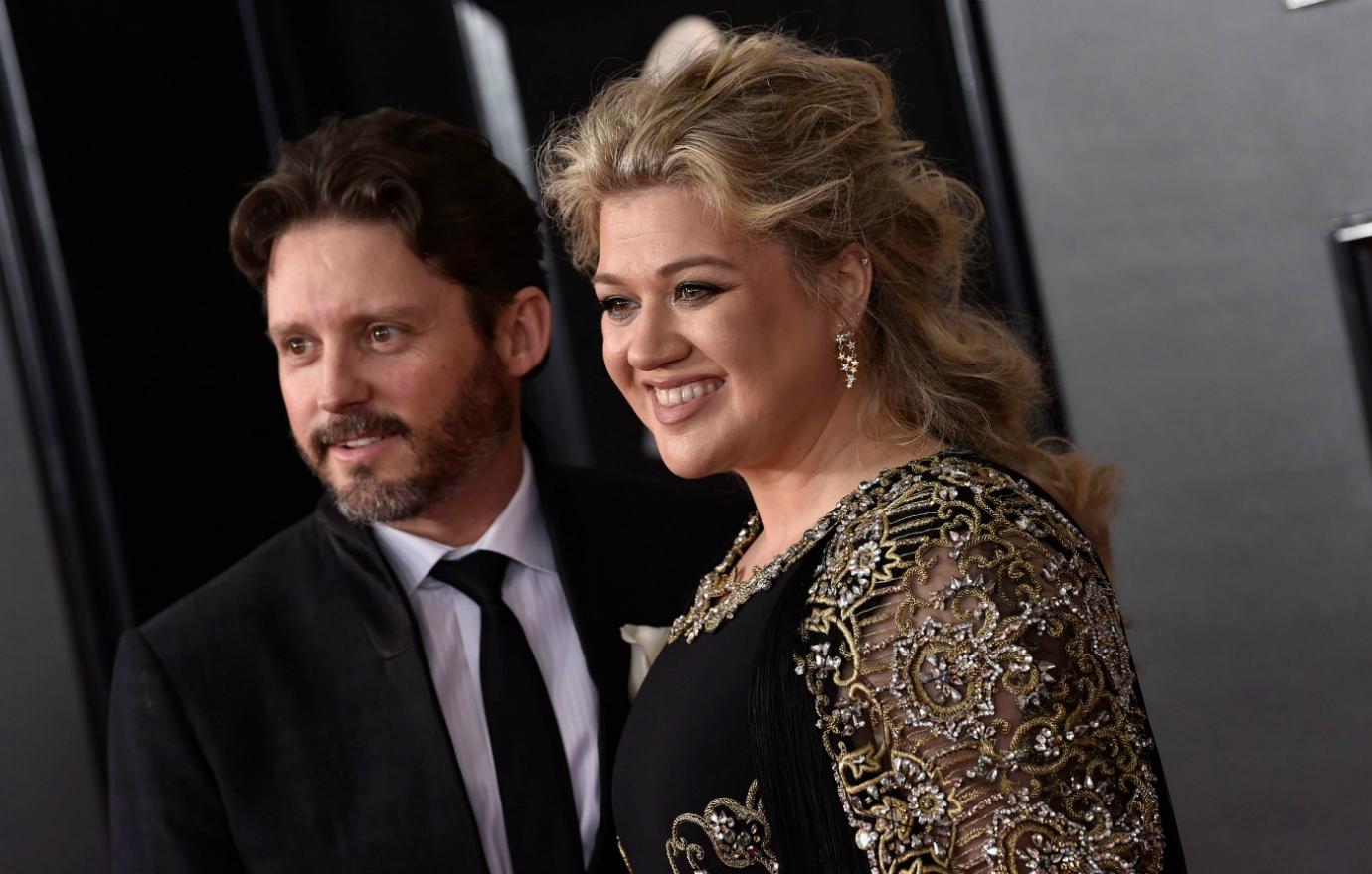 Kelly Clarkson, in a black patterned dress, stands on the red carpet next to Brandon Blackstock who wears a black suit and white tie.