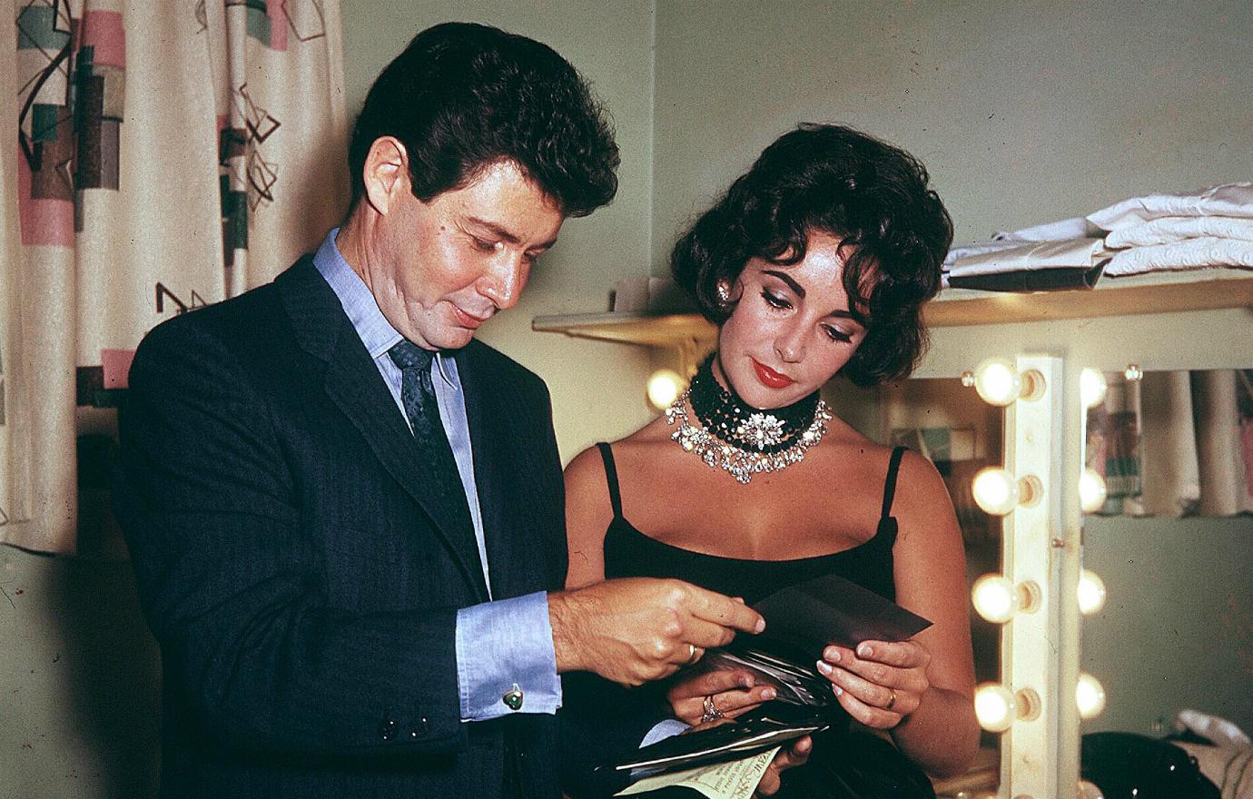 Eddie Fisher, in a black suit and blue tie, looks at photos alongside Elizabeth Taylor who wears a big black choker, a prominent diamond necklace, and a black dress.