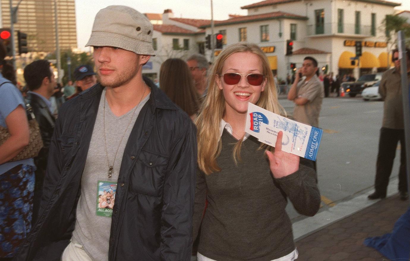 Ryan Phillippe, in a grey tshirt, hat, and black coat, walks alongside Reese Witherspoon who wears a grey sweater over a white shirt.