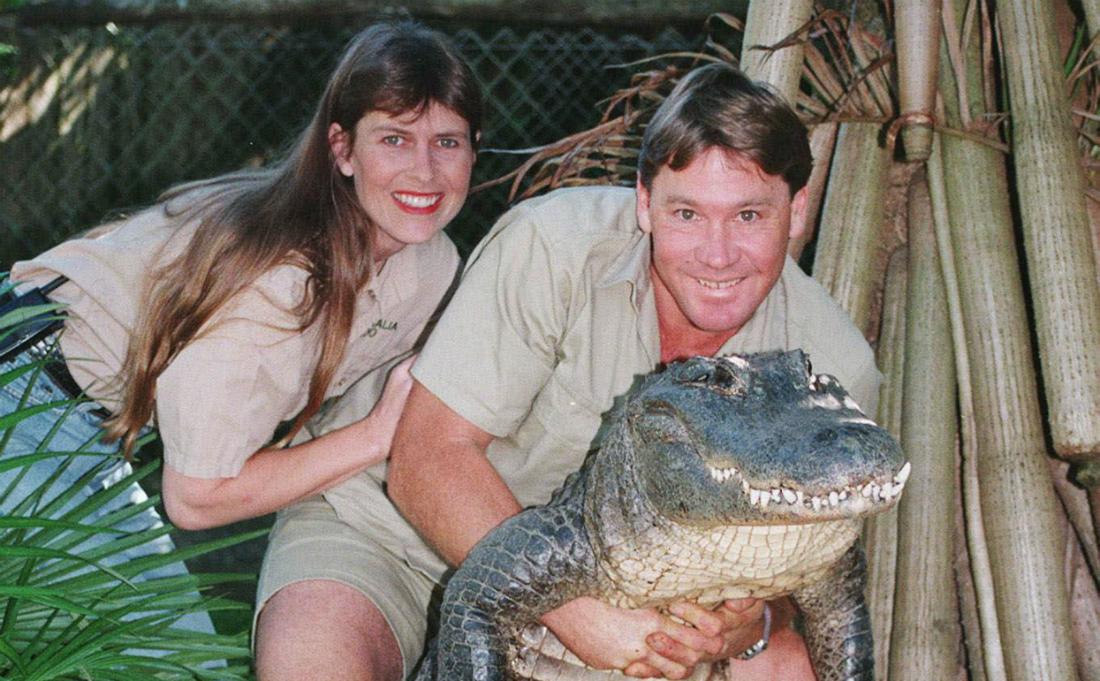 Dressed in khaki shorts and shirt, Steve Irwin smiles as he poses with an alligator. His wife, dressed in similar style, stands next to him, her hand on his arm.