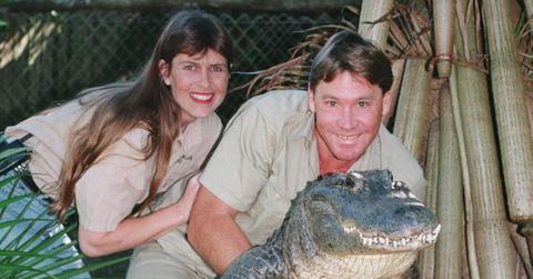 Dressed in khaki shorts and shirt, Steve Irwin smiles as he poses with an alligator. His wife, dressed in similar style, stands next to him, her hand on his arm.