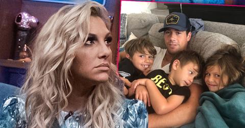 RHOC Gina Kirschenheiter Wearing Blue Print Dress Inset Husband Matt Wearing Baseball Cap Sitting On Couch With Their Three Kids