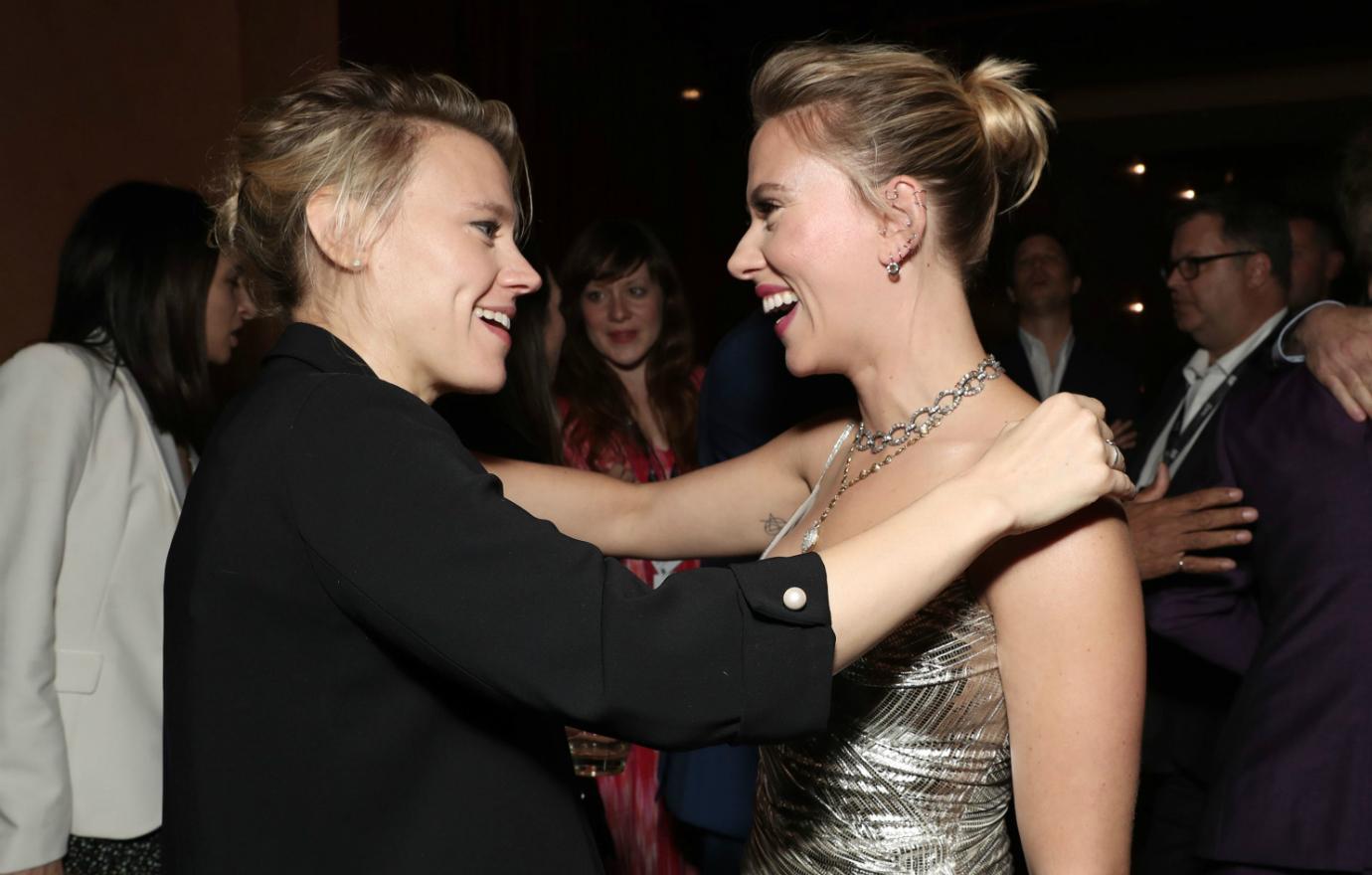 Scarlett Johansson has her arm on Kate McKinnon who has her arm on Scarlett.