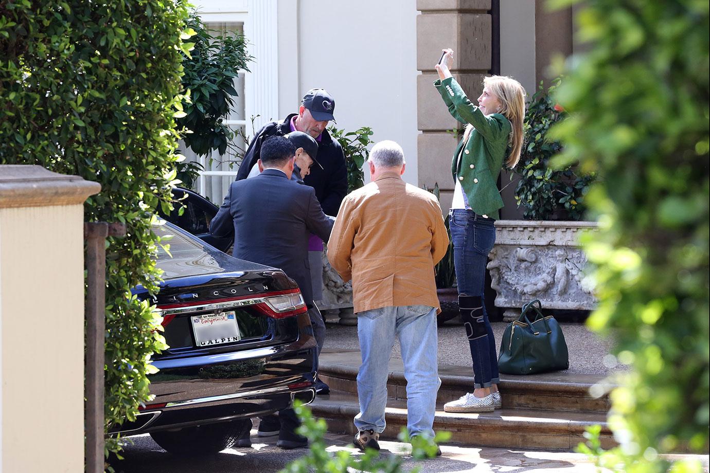 Larry King Wearing Baseball Cap Being Helped Out Of Car While Shawn Wearing a Green Jacket And Jeans Takes Photos of Trees With Phone