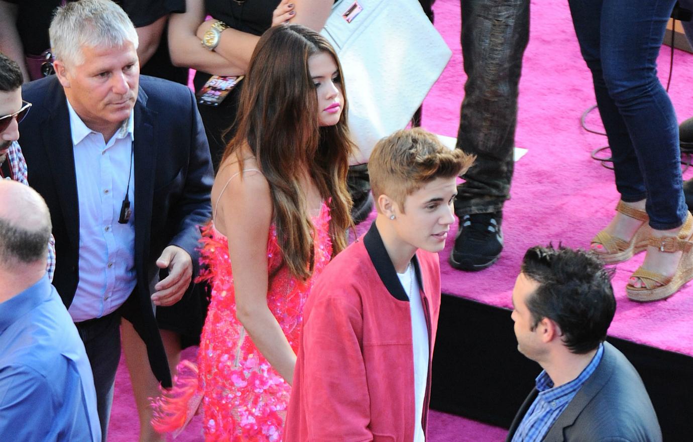 Justin Bieber, in a red jacket and white tshirt, leads Selena Gomez by the hand down a red carpet. She's also in red.