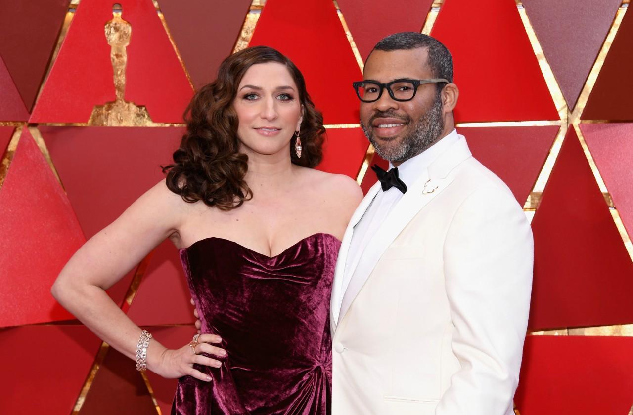 Jordan Peele in a white suit and Chelsea Peretti in a red gown, stand in front of a red background