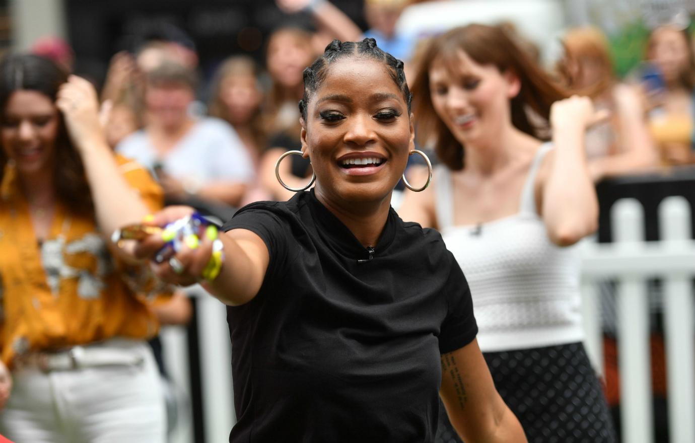 Keke Palmer in a black top and big hoop earrings smiles and points.