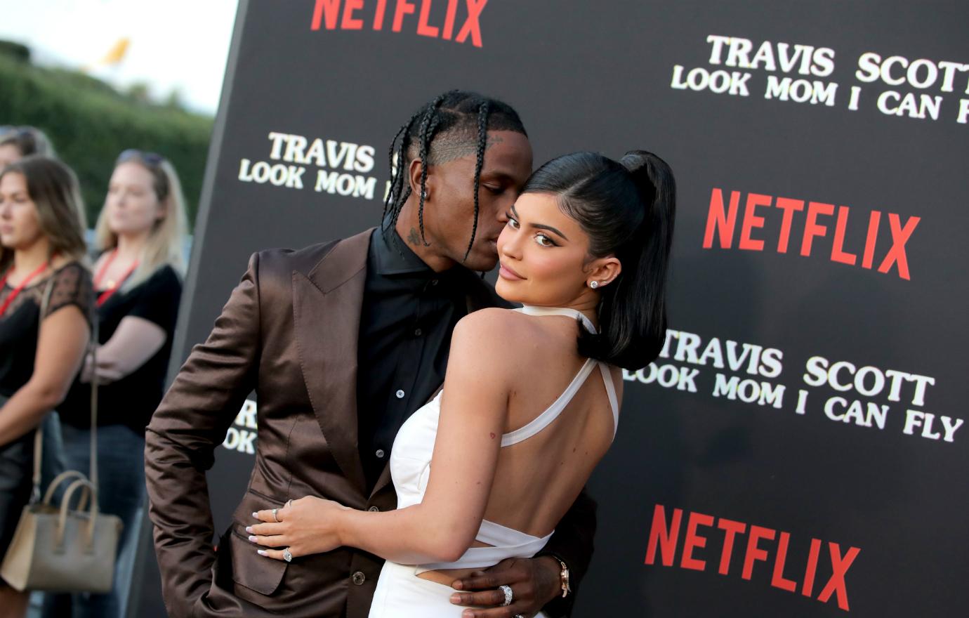 Travis Scott, in a chocolate brown suit with black tie, kisses Kylie Jenner, who wears a white dress, on the red carpet.