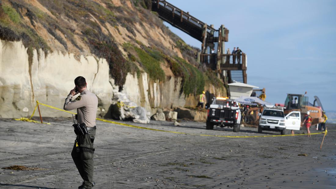 A San Diego County Sheriff's deputy looks on as search and rescue personnel work at the site of a cliff collapse at a popular beach, in Encinitas, Calif.