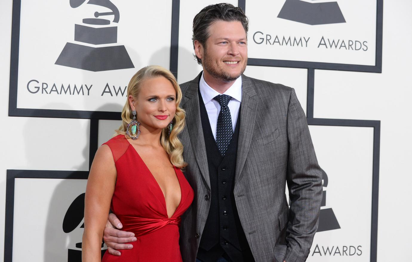 Miranda Lambert, in a red dress, stands next to Blake Shelton who wears a three piece grey suit.