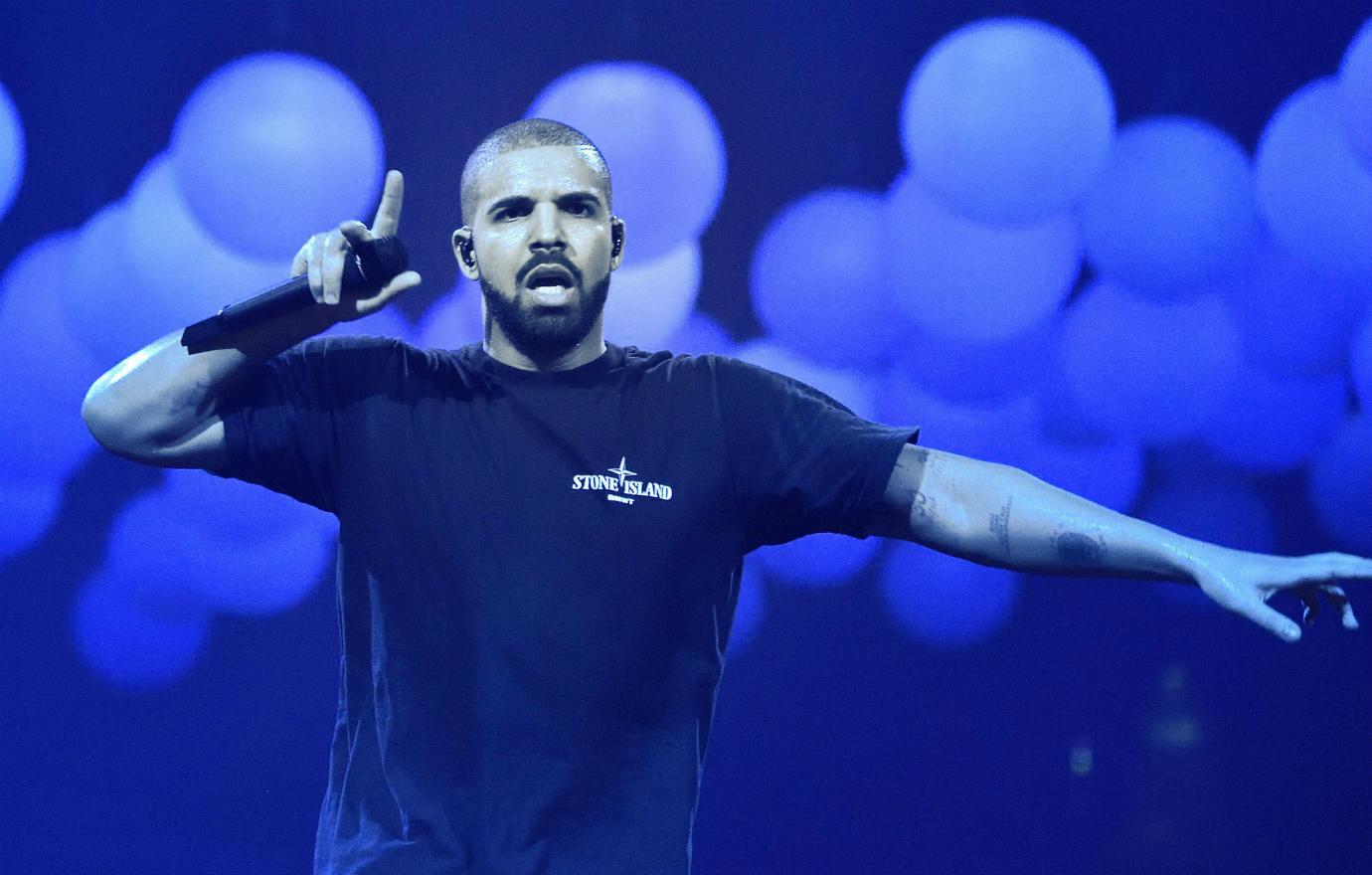 Drake stands on stage, holding a mic, wearing a top with a Stone Island logo.