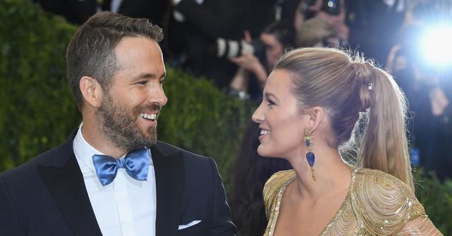 Ryan Reynolds in a black suit and Blake Lively in a gold gown smile at one another one the red carpet with paparazzi in the background