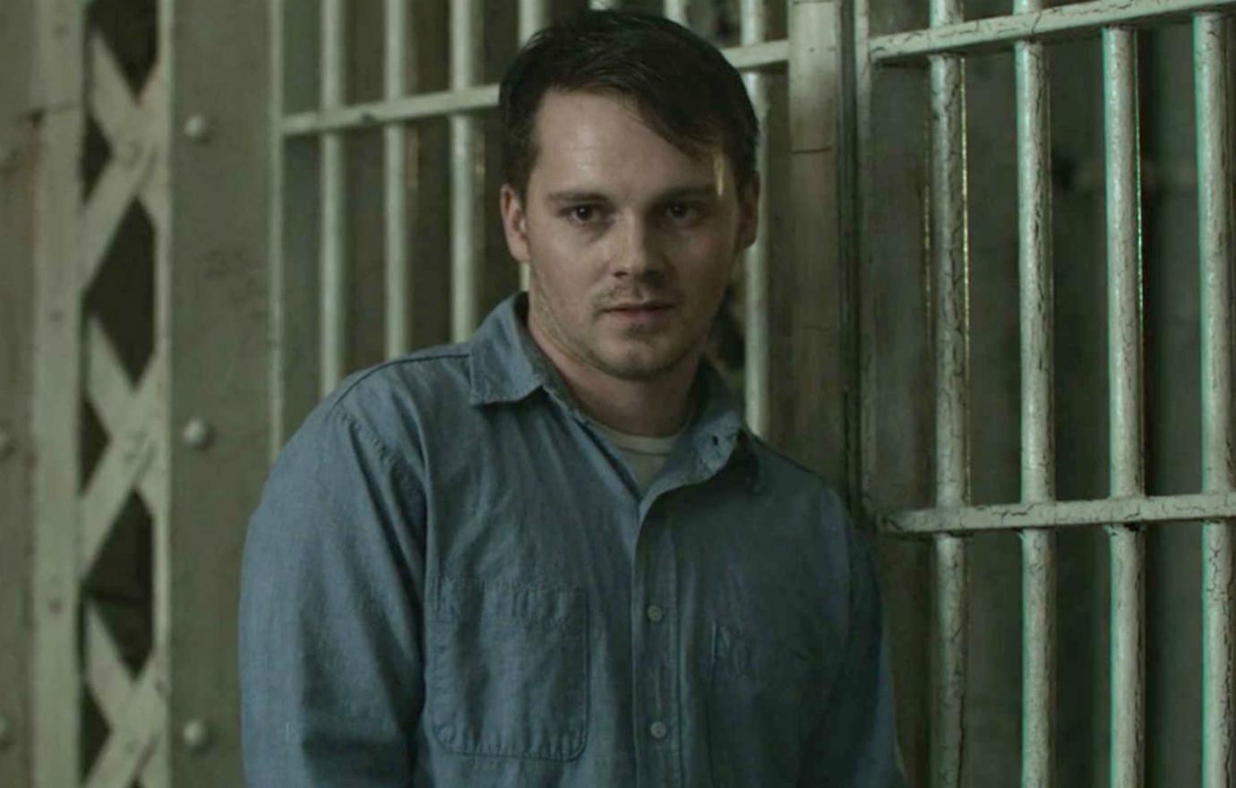 Sam Strike stands near his prison cell in his prison blues in a scene from Mindhunter where he portrays killer Monte Ralph Rissell.