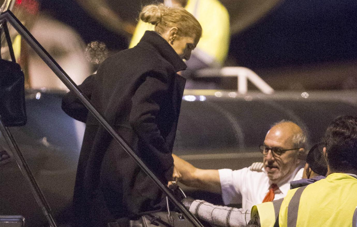 Celine Dion clad in a black jacket with high collar walks down the gangplank of her private jet while airport workers wait below for her.