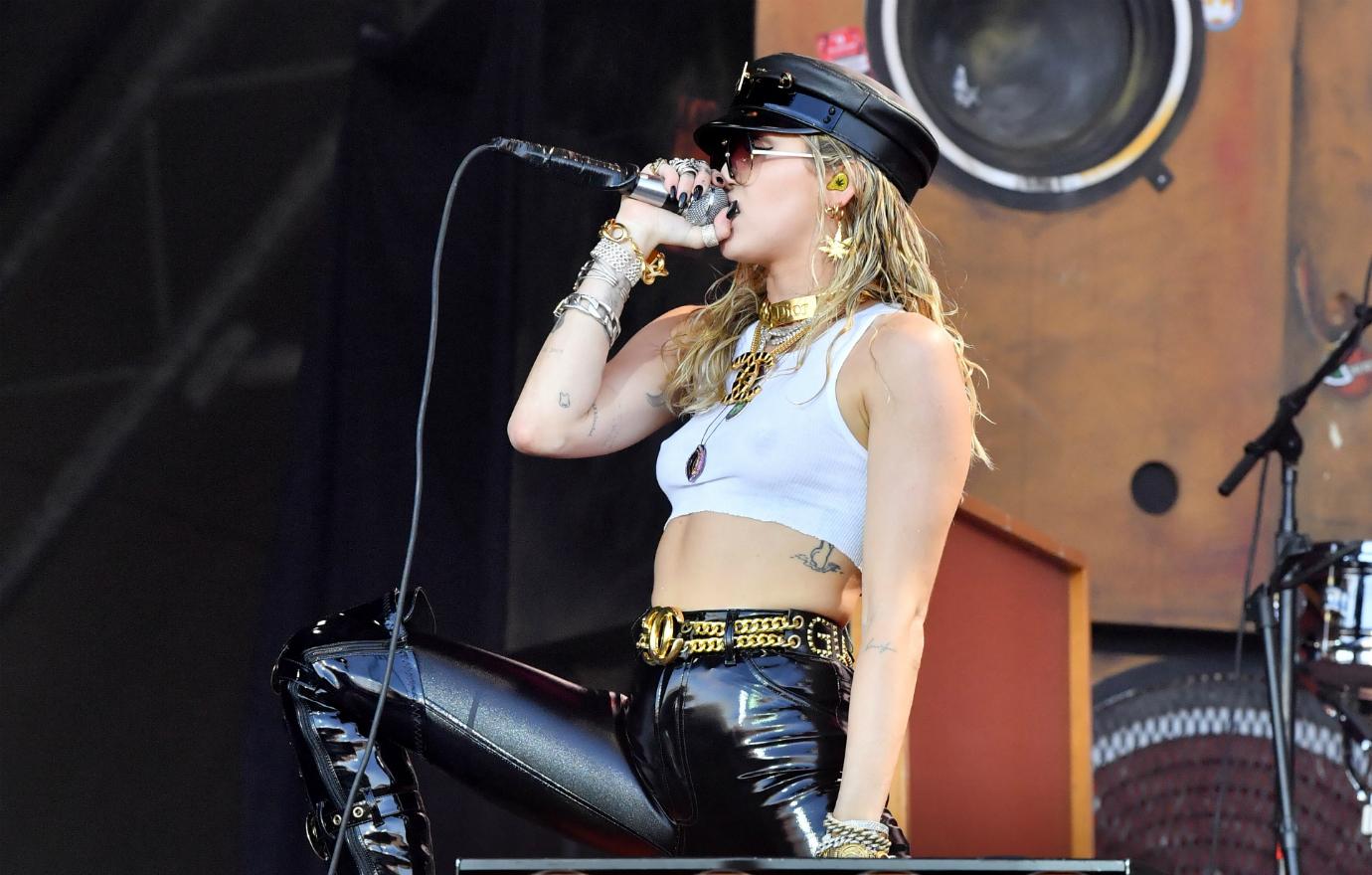 Miley Cyrus performs on stage in leather pants, cropped top, and black hat.