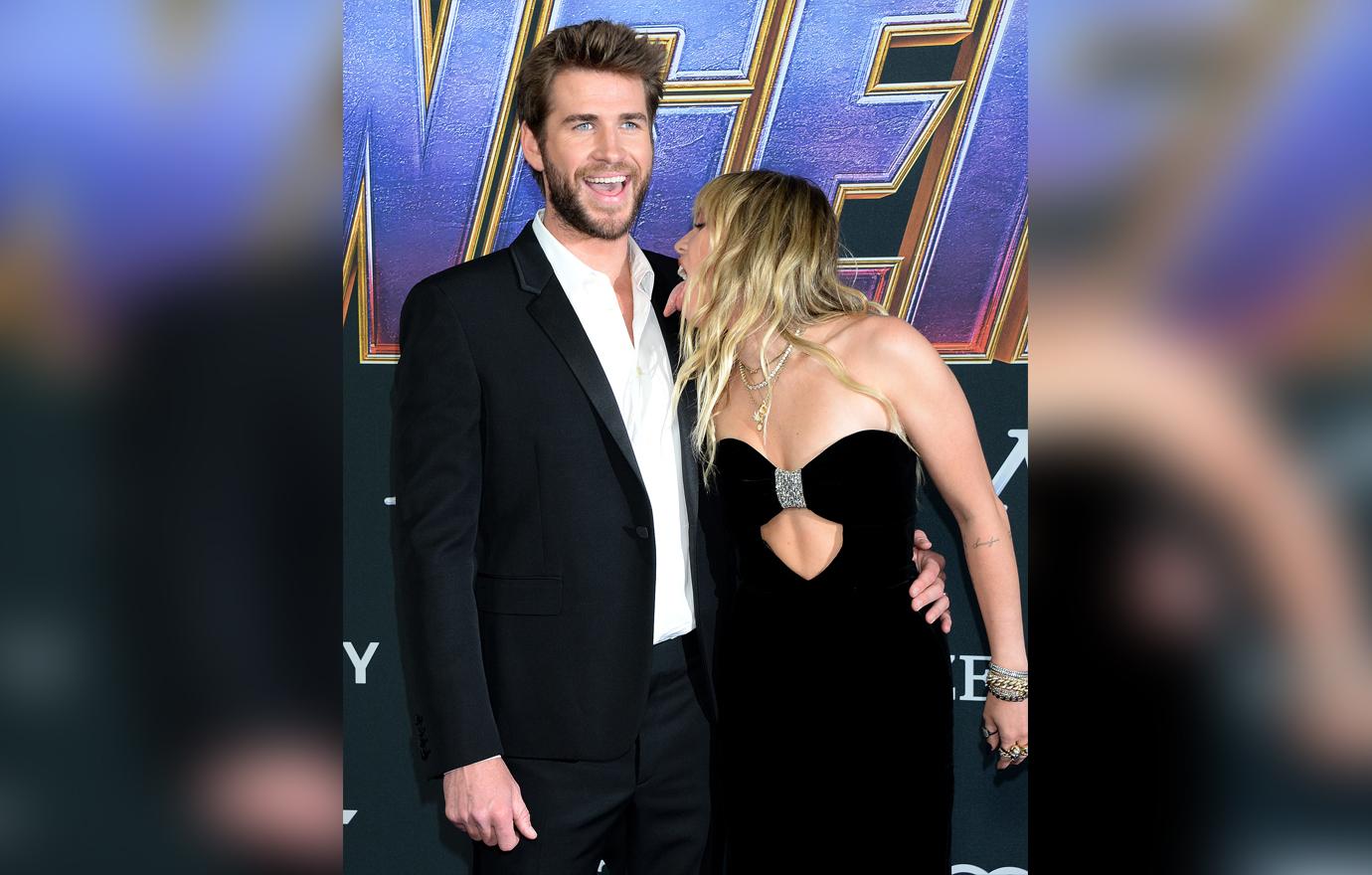 Liam Hemsworth Laughs Wearing a Suit on The Avengers Premiere Red Carpet As Miley Cyrus Poses Licking Towards His Neck Wearing A Tight Black Dress With Cut Out