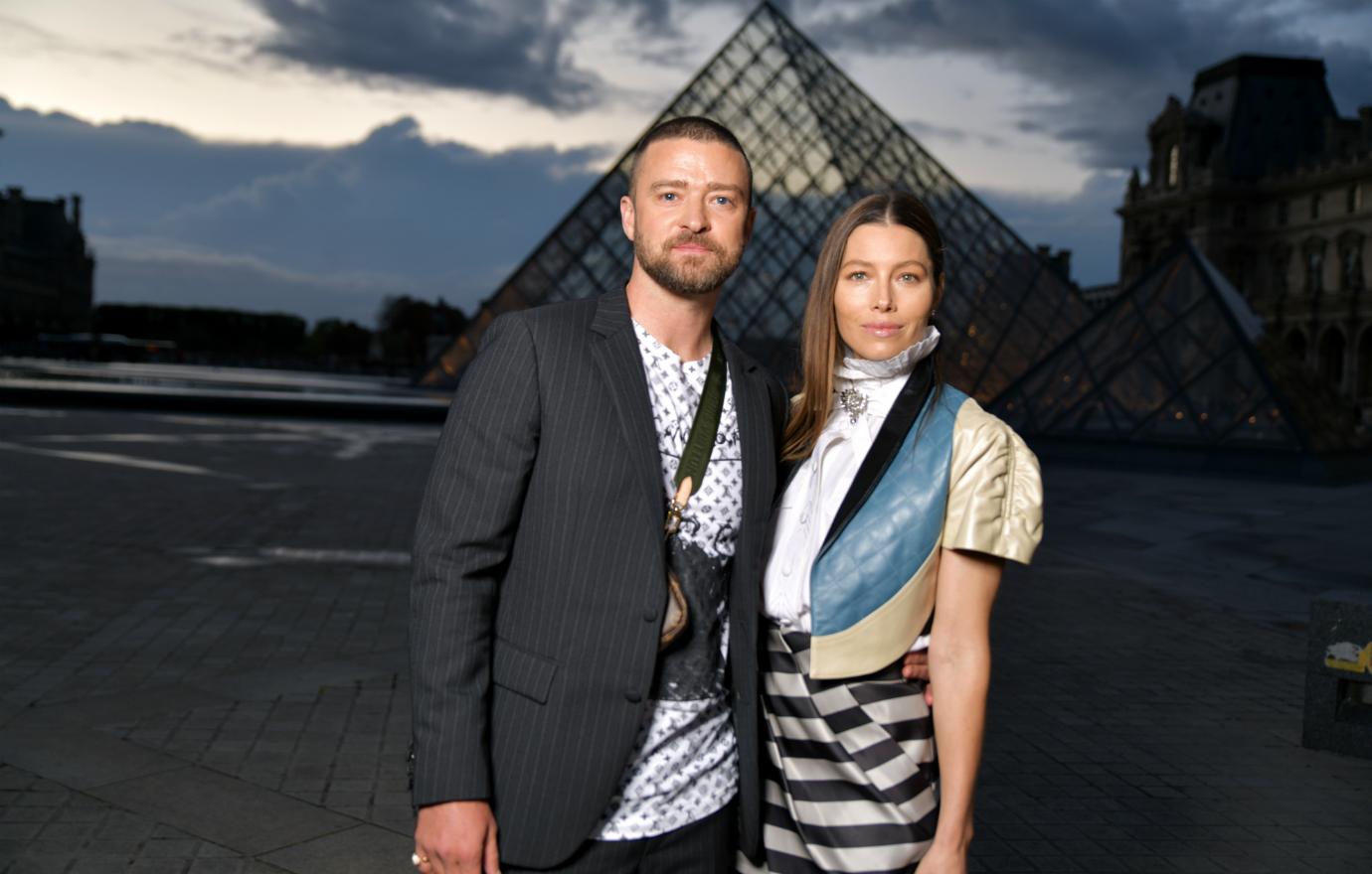 Justin Timberlake, in a black jacket and patterned tshirt, stands next to Jessica Biel who wears a multi-colored, multi-patterned dress and jacket and a white blouse.