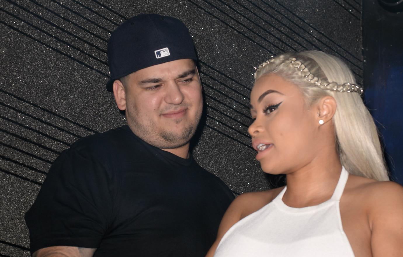 ROb Kardashian, in a black shirt and backwards baseball cap looks at Blac Chyna who wears a white dress.