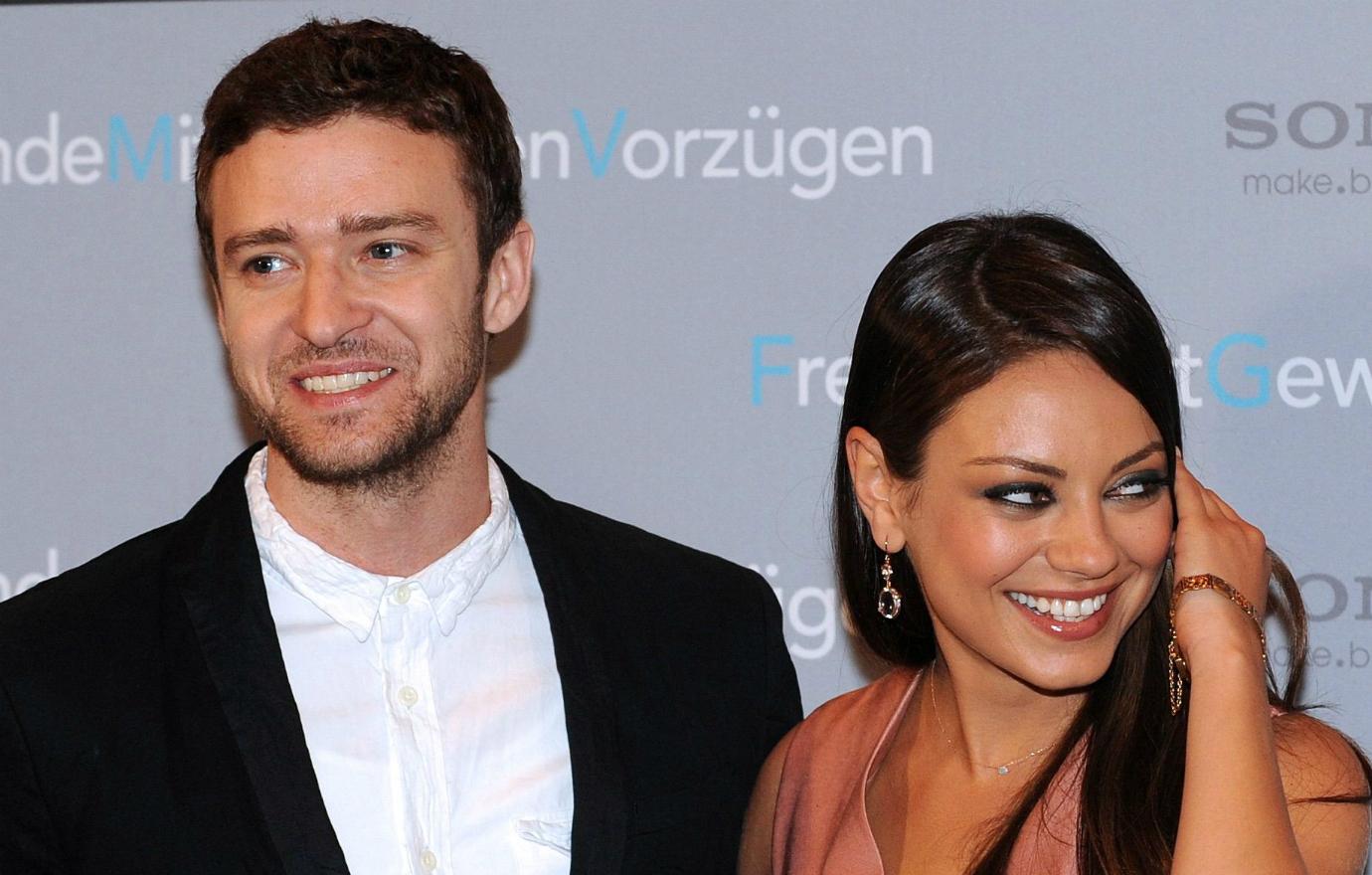 Justin Timberlake stands on the red carpet in white shirt and black jacket slongside Mila Kunis who wears a peach-colored dress.