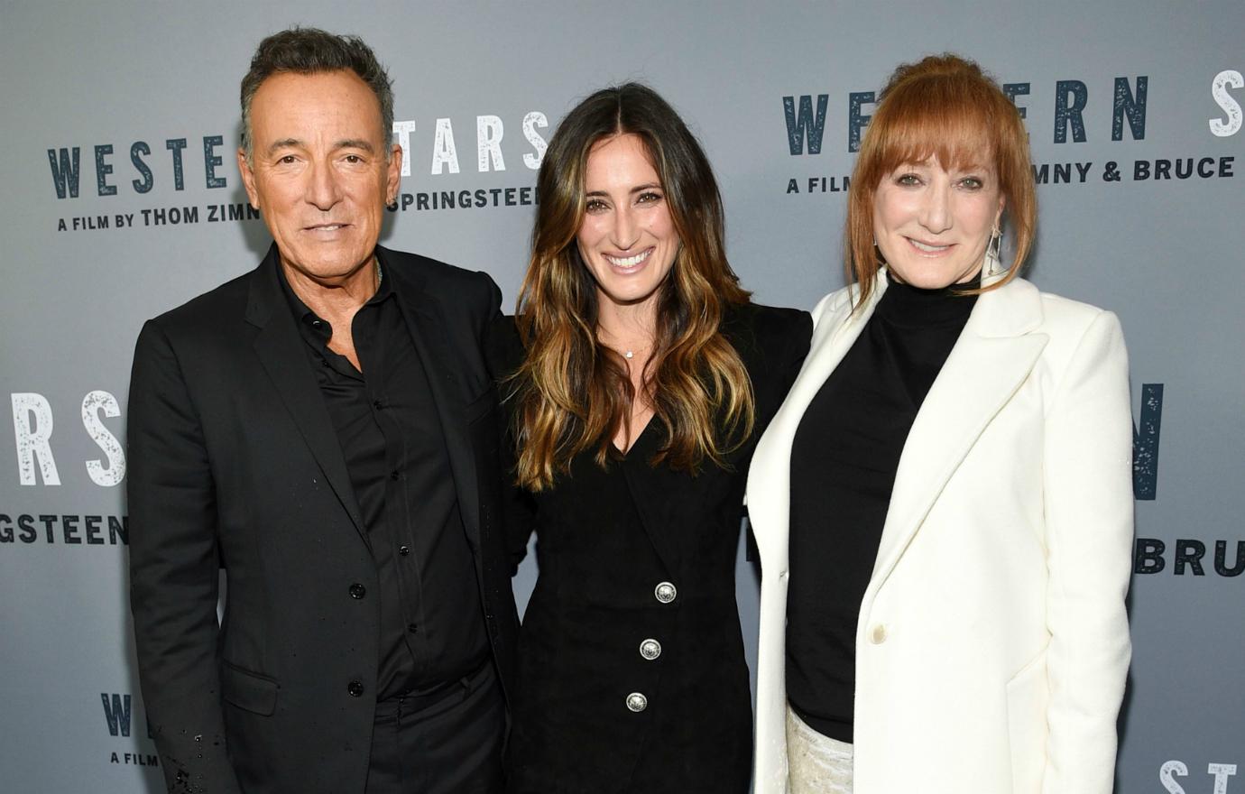 Bruce Springsteen, in a black suit, stands next to daughter Jessica who wears a black dress, and wife Patti Scialfa, who wears a white coat over a black top and white pants.