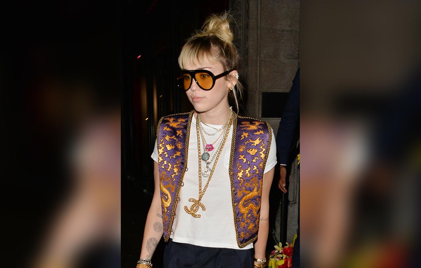Miley Cyrus Looks Serious as She Walks Outside a Restaurant Wearing Large Orange Sunglasses a White Top With A Orange and Purple Vest and Large Chanel Chain Necklaces