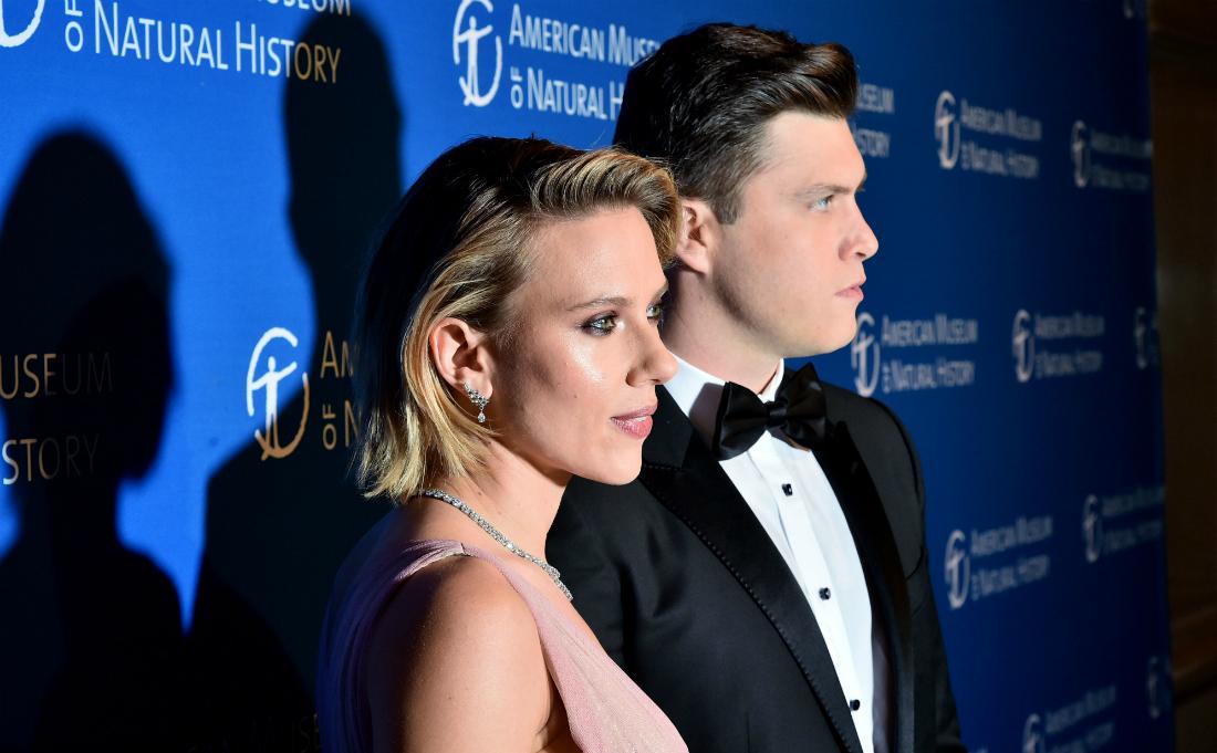 Colin Jost, in a black tux, stands next to Scarlett Johansson who wears a pink dress on the red carpet.