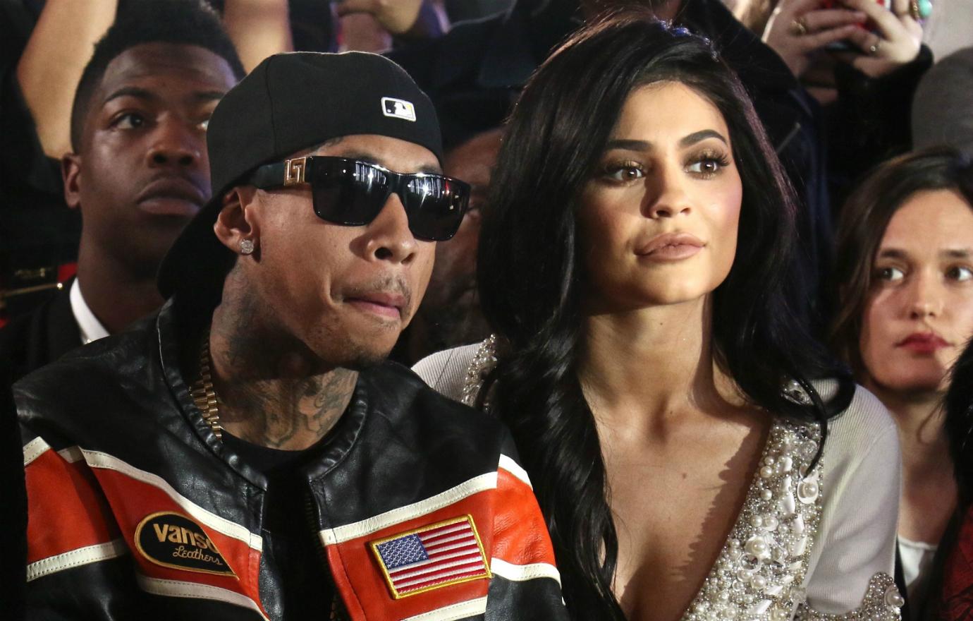 Tyga, in shades, baseball cap, and black leather jacket sits next to Kylie Jenner who wears a grey patterned dress.