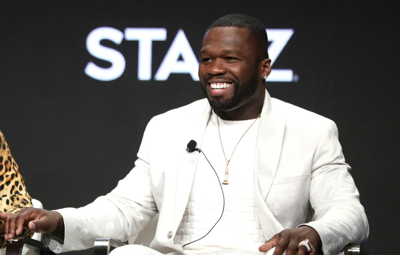 50 Cent sits on stage in an all-white outfit, smiling.