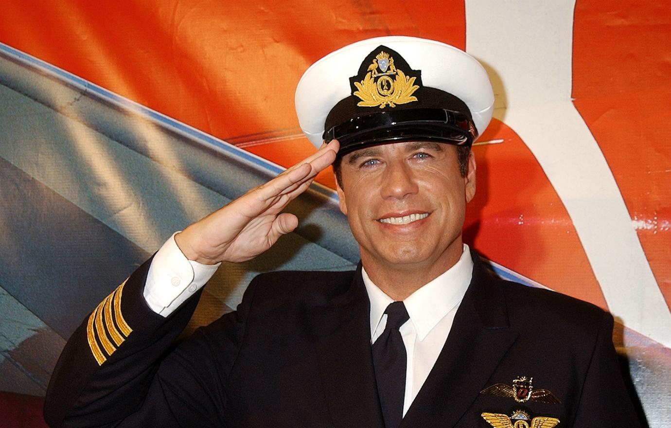 John Travolta, in pilot’s uniform, salutes and smiles.