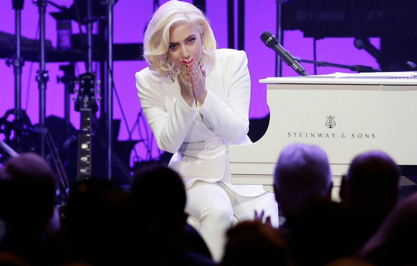 Lady Gaga, in white, sits at a white piano.