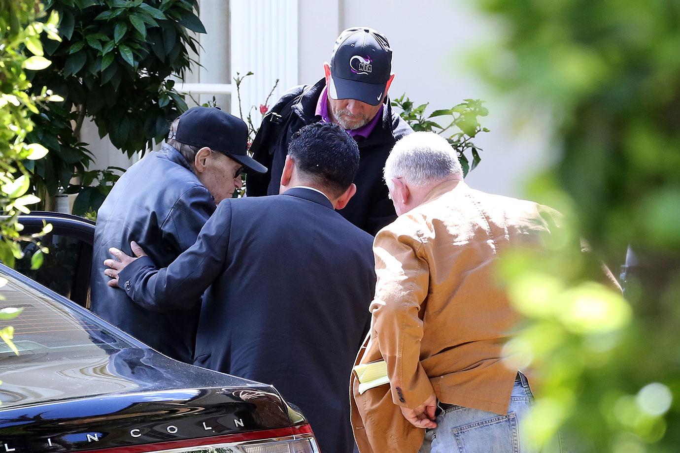 Larry King Wearing Baseball Cap Being Helped Out Of Car