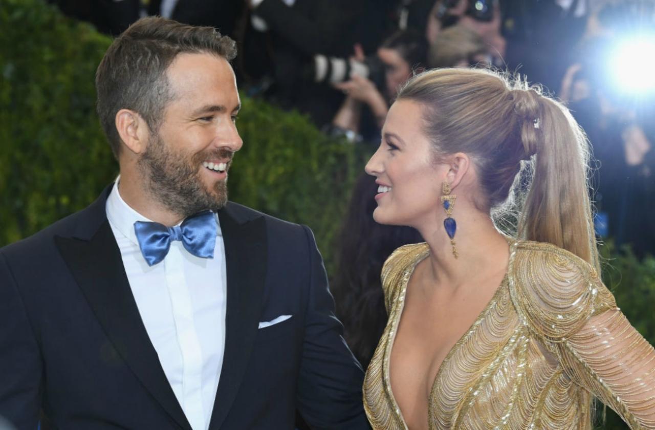 Ryan Reynolds in a black suit and Blake Lively in a gold gown smile at one another on the red carpet