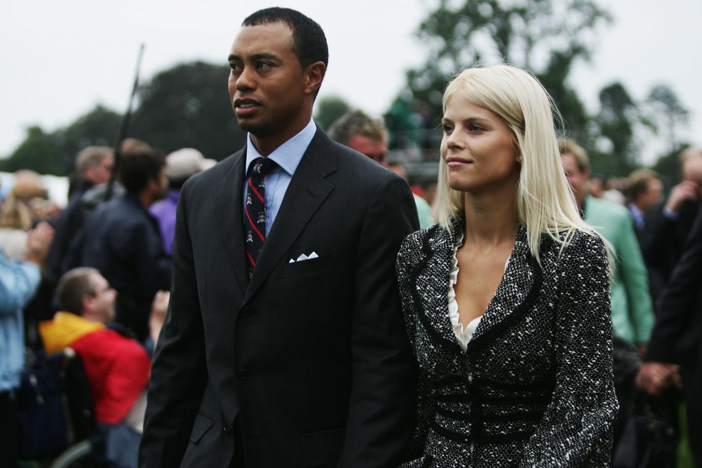 Elin Nordegren wears a grey suit jacket while standing next to tiger woods who sports a suit.