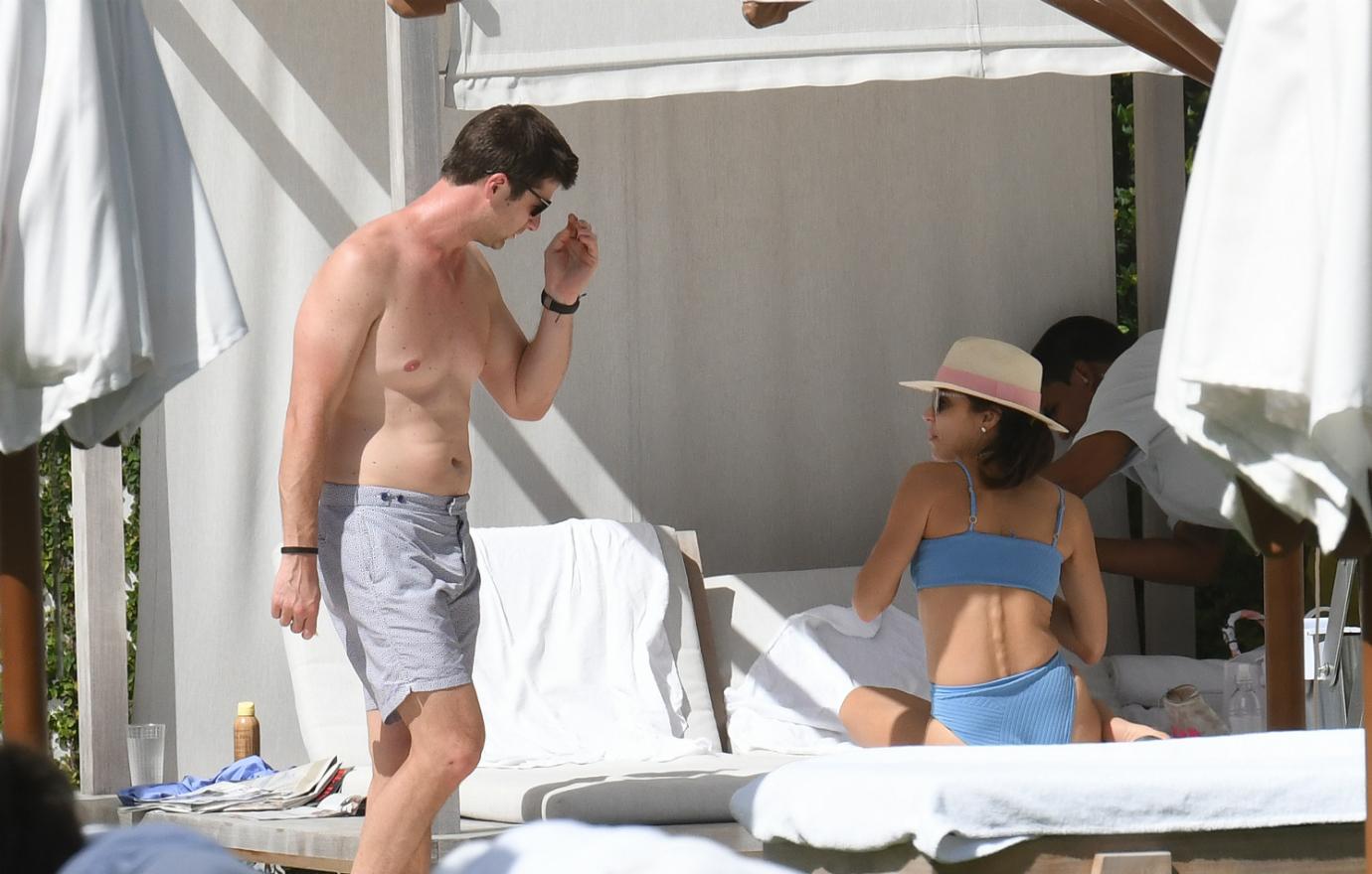 Paul bernon, in just shorts, stands by Bethenny Frankel who sits on a lounger in a blue bikini and hat.