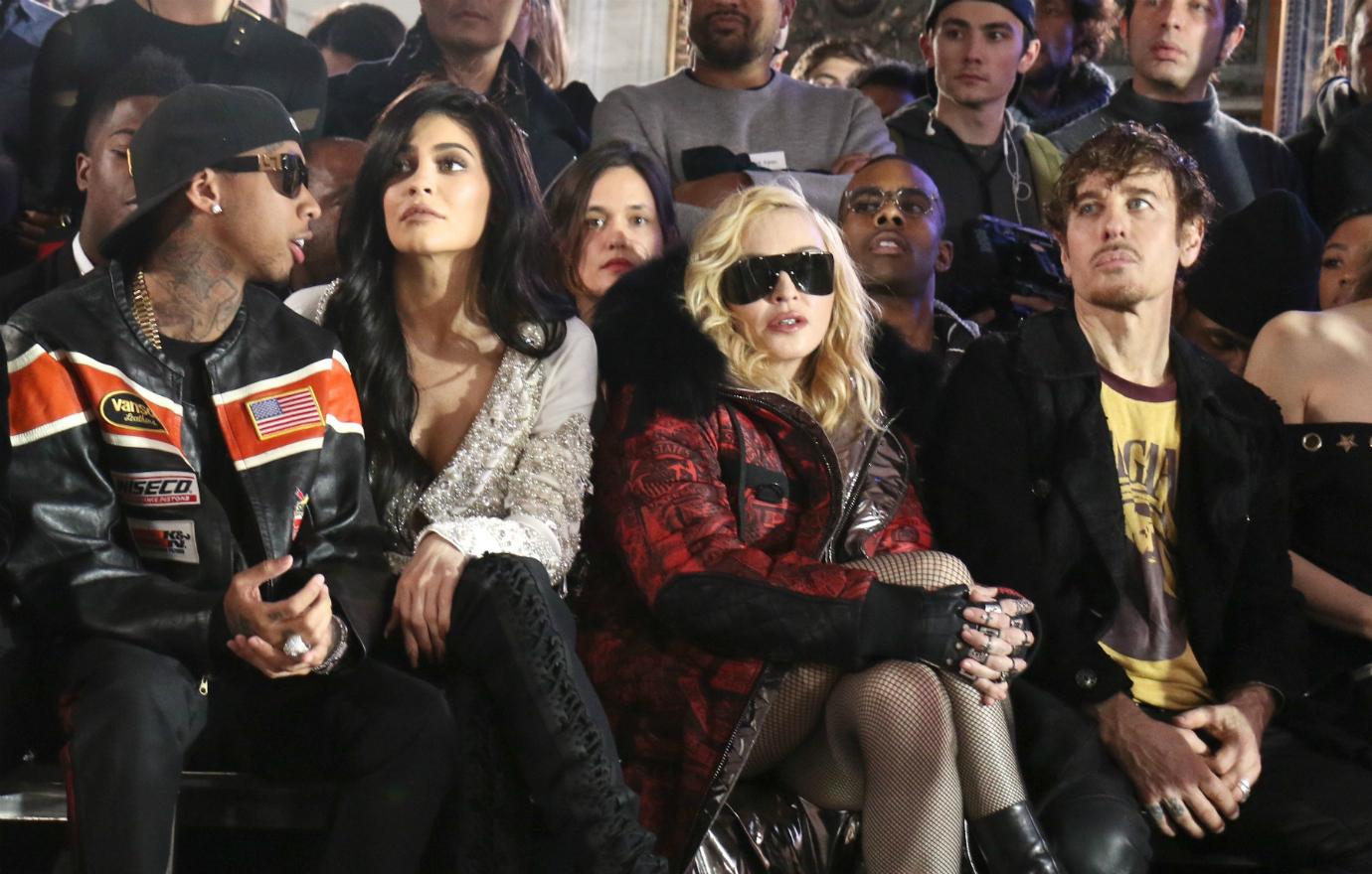 Tyga wears a baseball hat, shades, black leather jacket with a red stripe, and black jeans. He sits next to Kylie Jenner who wears black boots, black pants, and a white patterned top.