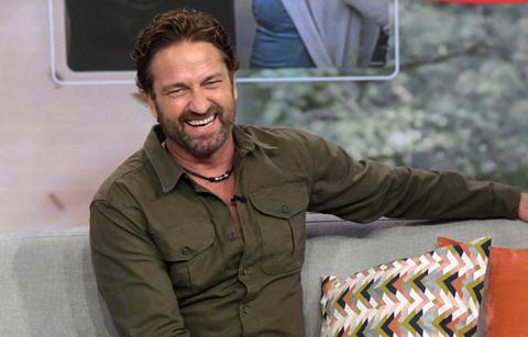 Gerard Butler smiled during an interview wearing a casual brown buttoned-down shirt and black slacks.