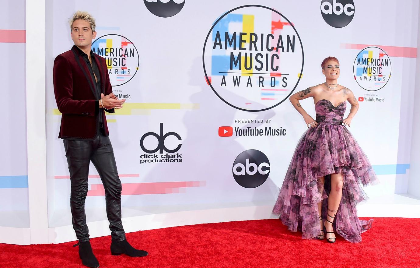 Halsey, in a pink patterned dress, stands on the red carpet a few feet away from G-Eazy who ears a dark suit.