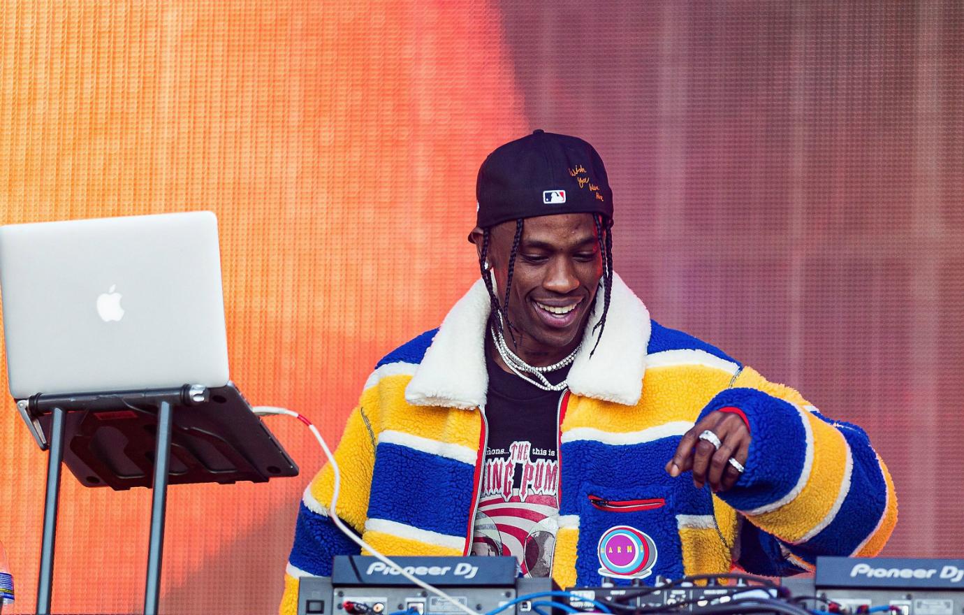 Travis Scott on stage in a baseball cap, yellow/white/blue striped jacket and tshirt.