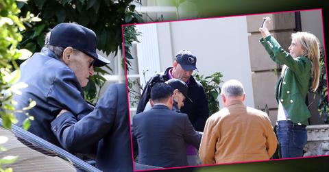 Larry King Wearing Baseball Cap Being Helped Out Of Car While Shawn Wearing a Green Jacket And Jeans Takes Photos of Trees With Phone